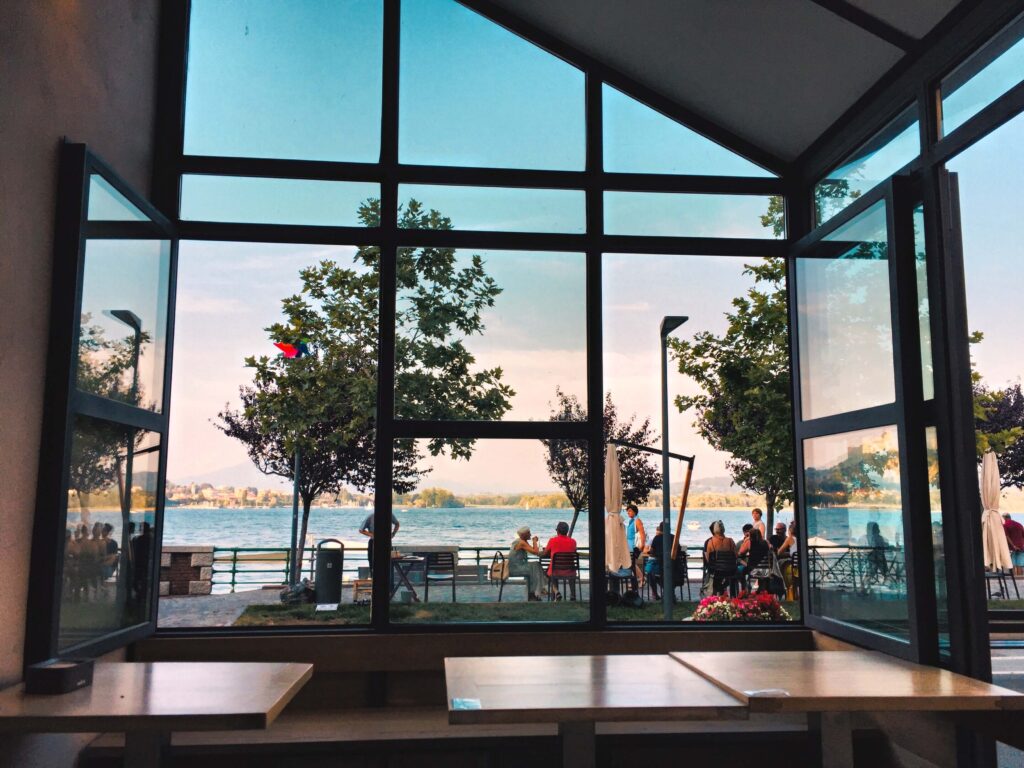 Photo of a the interior of a restaurant with floor extensive windows that look out on to a body of water with a small group of people sitting outside the restaurant